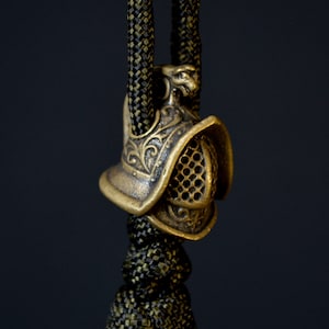 May include: A gold-colored metal charm in the shape of a gladiator helmet with intricate details. The helmet has a raised design and a mesh face guard. The charm is attached to a black and green cord.