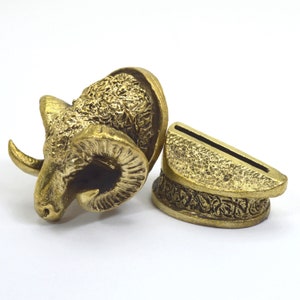 Op de afbeelding: Een gouden metalen rammenkopfiguur met ingewikkelde details en een bijpassende standaard. De rammenkop is naar links gericht en heeft een gedetailleerd gezicht en hoorns. De standaard is een eenvoudige rechthoekige vorm met een gleuf aan de bovenkant.