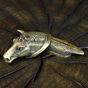 May include: A gold-toned metal horse head bottle stopper with a detailed design. The stopper is on a dark brown wooden surface.