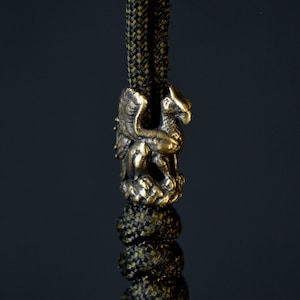 May include: A gold-coloured metal charm in the shape of a griffin, with intricate details, attached to a green and black paracord.