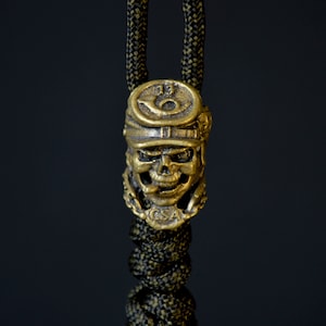 May include: A gold-colored skull charm with a military-style hat and the text "13 CSA" on the front. The charm is attached to a black and green cord.
