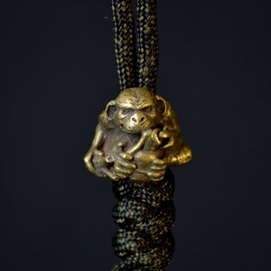 Pode incluir: Um amuleto de macaco dourado com um design detalhado, pendurado em um cordão preto e verde.