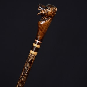 Pode incluir: Uma bengala de madeira com uma alça esculpida em forma de cabeça de urso. A bengala é tingida de marrom escuro e tem detalhes em cobre.