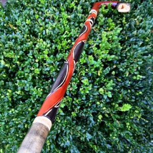 May include: A wooden walking stick with a red, black and white design. The stick is carved with intricate patterns and has a smooth, polished finish.