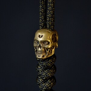 Könnte beinhalten: Eine goldene Schädelperle mit filigranen Details, befestigt an einem grün-schwarzen Paracord.