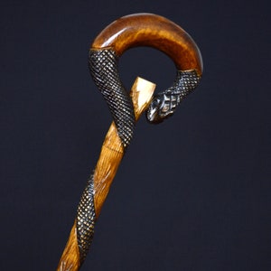 Puede incluir: Un bast&oacute;n de madera con un mango tallado en forma de cabeza de serpiente. La serpiente est&aacute; envuelta alrededor del mango y su cola est&aacute; enroscada. El bast&oacute;n est&aacute; hecho de madera marr&oacute;n claro y la serpiente est&aacute; pintada de negro.