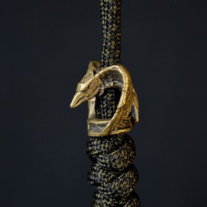 Puede incluir: Una cuenta de metal de color dorado con un diseño detallado de cabeza de dragón. La cuenta está unida a un cordón de paracord negro y verde.