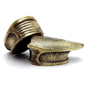 May include: Two antique brass-colored rings with textured surfaces and intricate designs. One ring is positioned on top of the other, showcasing the detailed craftsmanship. The rings have a weathered appearance, suggesting age and use.