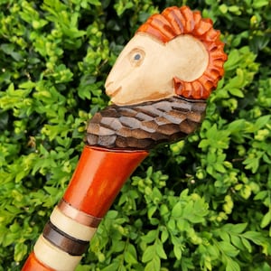 May include: A wooden walking stick with a carved ram's head at the top. The ram's head is painted orange and brown, and the stick is painted orange and brown with white and black accents.
