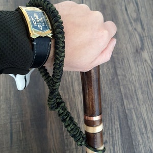 Puede incluir: Una pulsera de paracord verde y negro con un reloj dorado y un bast&oacute;n de madera marr&oacute;n con detalles en cobre y blanco.