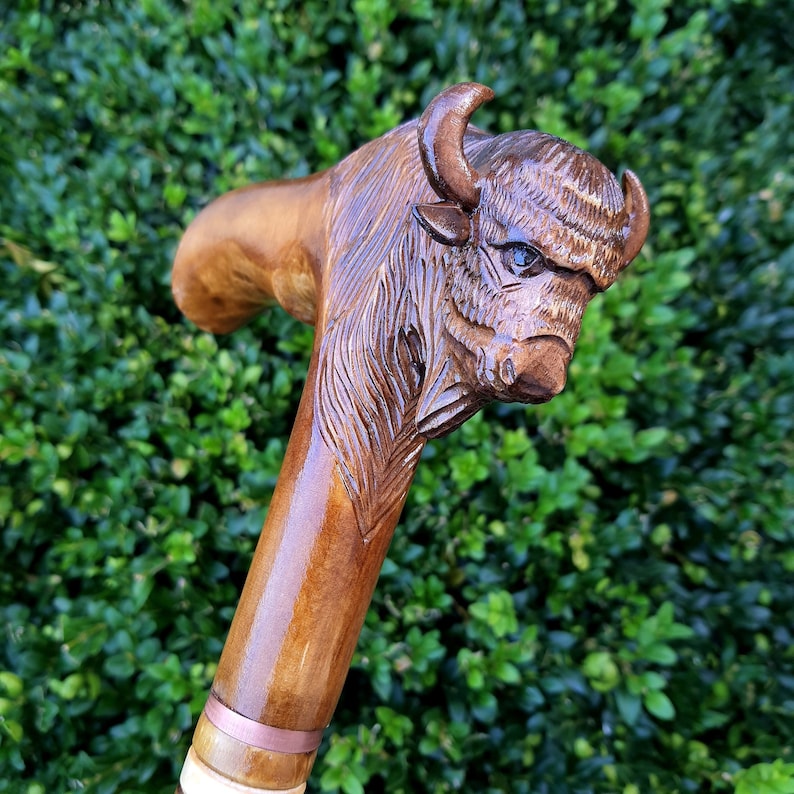 May include: A wooden walking stick with a carved bison head. The bison is brown and has a detailed face with a textured fur pattern. The stick is polished and has a copper band near the bottom.