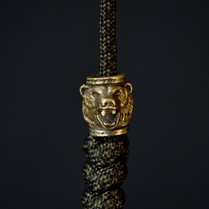 Könnte beinhalten: Ein goldfarbener Bärenkopf-Anhänger mit detailliertem Design, befestigt an einem schwarzen und goldenen Paracord.