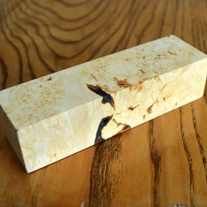 Könnte beinhalten: Ein Holzblock mit einem einzigartigen Maserungsmuster. Das Holz ist hellbraun mit dunkelbraunen Streifen und Wirbeln. Der Block ist rechteckig und hat glatte Kanten.