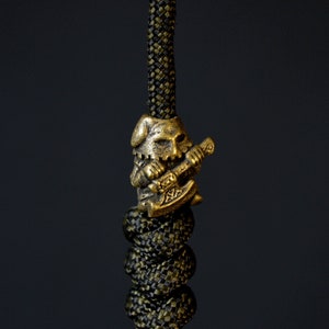 May include: A gold-colored metal bead with a detailed design of a warrior holding an axe. The bead is attached to a black and green paracord.