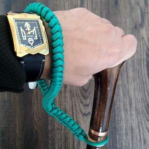 Puede incluir: Una pulsera de paracord verde con un reloj dorado con una banda negra y una esfera dorada con un s&iacute;mbolo de tridente y la palabra "VERNIVA" en ella. La pulsera est&aacute; unida a un bast&oacute;n de madera marr&oacute;n con una banda de cobre.