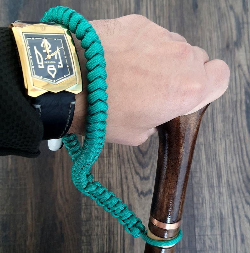 Puede incluir: Una pulsera de paracord verde con un reloj dorado con una banda negra y una esfera dorada con un s&iacute;mbolo de tridente y la palabra "VERNIVA" en ella. La pulsera est&aacute; unida a un bast&oacute;n de madera marr&oacute;n con una banda de cobre.