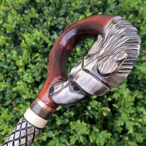 Könnte beinhalten: Ein Gehstock aus Holz mit einem geschnitzten Wolfskopf als Griff. Der Griff ist dunkelrot gebeizt und das Gesicht des Wolfes ist in Grautönen bemalt. Der Stock ist mit einem Kupferband und einem weißen und schwarzen Muster verziert.