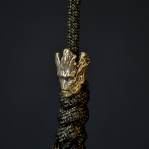 May include: A gold-coloured metal charm featuring a detailed depiction of the character Groot from the Guardians of the Galaxy franchise. The charm is attached to a green and black paracord.