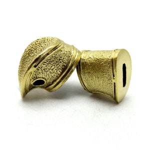 Könnte beinhalten: Zwei goldfarbene Schwertknauf aus Metall mit einer strukturierten Oberfläche. Ein Knauf hat eine runde Öffnung, während der andere ein komplizierteres Design mit einer kleineren Öffnung hat.