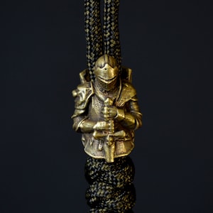 Pode incluir: Um amuleto de metal dourado de um cavaleiro com armadura segurando uma espada. O amuleto está preso a um cordão de paracord preto e verde.
