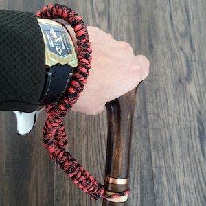Puede incluir: Un brazalete de paracord negro y rojo con un reloj dorado y un bast&oacute;n de madera con una banda de cobre.