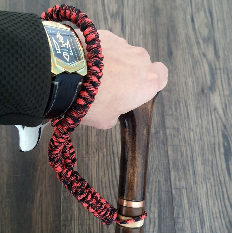 Puede incluir: Un brazalete de paracord negro y rojo con un reloj dorado y un bast&oacute;n de madera con una banda de cobre.
