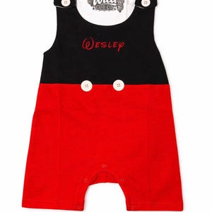 Puede incluir: Un pelele de bebé rojo y negro con botones blancos y el nombre "Wesley" bordado en rojo. El pelele tiene un cuello blanco con el texto "Wild Cub Co" y gráficos de osos. El pelele sin mangas tiene broches en la parte inferior.