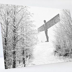 May include: A black and white photograph of the Angel of the North sculpture in a snowy landscape. The sculpture is a large, winged figure standing on a hill. The trees in the foreground are covered in snow.