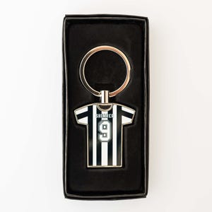 May include: A black and white striped jersey-shaped keychain with the name "SHEARER" and the number "9" on it. The keychain is in a black gift box with a gold-colored key ring.