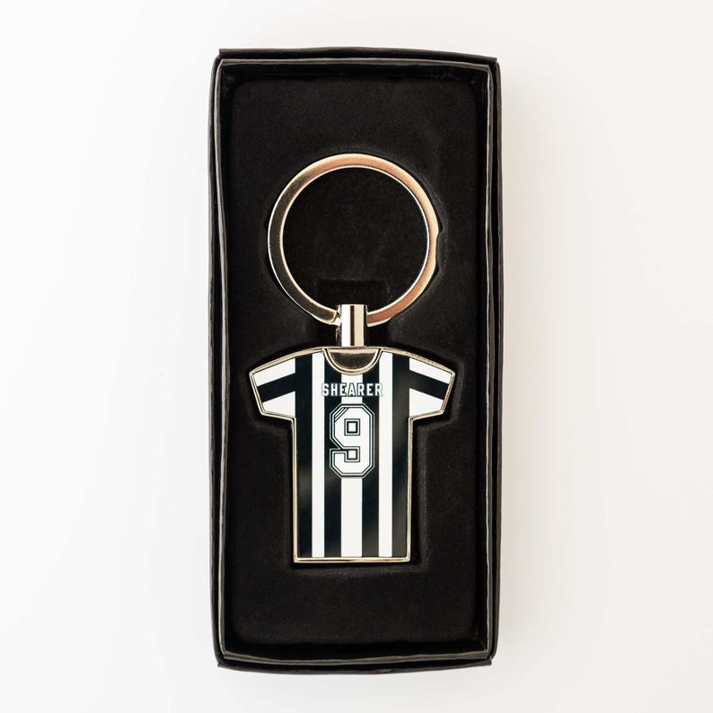 May include: A black and white striped jersey-shaped keychain with the name "SHEARER" and the number "9" on it. The keychain is in a black gift box with a gold-colored key ring.