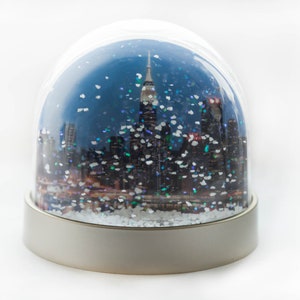 Könnte beinhalten: Eine gläserne Schneekugel mit einer Stadtlandschaft im Inneren. Die Stadt ist vor einem blauen Himmel mit fallendem weißen Glitzer silhouettiert. Der Sockel der Schneekugel ist silberfarben.