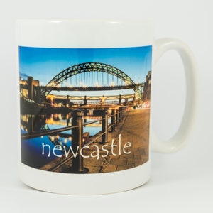 Puede incluir: Taza de cerámica blanca con una foto del Puente Tyne en Newcastle upon Tyne, Inglaterra, de noche. El puente está iluminado y se refleja en el agua. El texto "newcastle" está impreso en la taza.