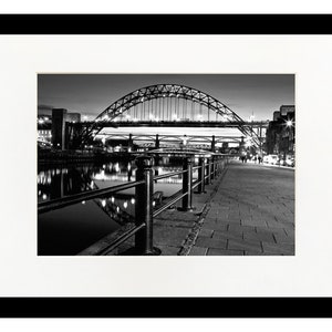 Puede incluir: Una fotografía en blanco y negro de un puente sobre un río de noche. El puente está iluminado con luces, y el agua refleja las luces. Hay una acera a lo largo del río, y una barandilla a lo largo del borde de la acera.