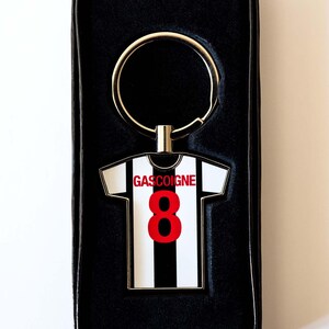 Puede incluir: Llavero con forma de camiseta de f&uacute;tbol a rayas blancas y negras con el nombre "GASCOIGNE" y el n&uacute;mero "8" en rojo. Presentado en una caja de regalo negra.
