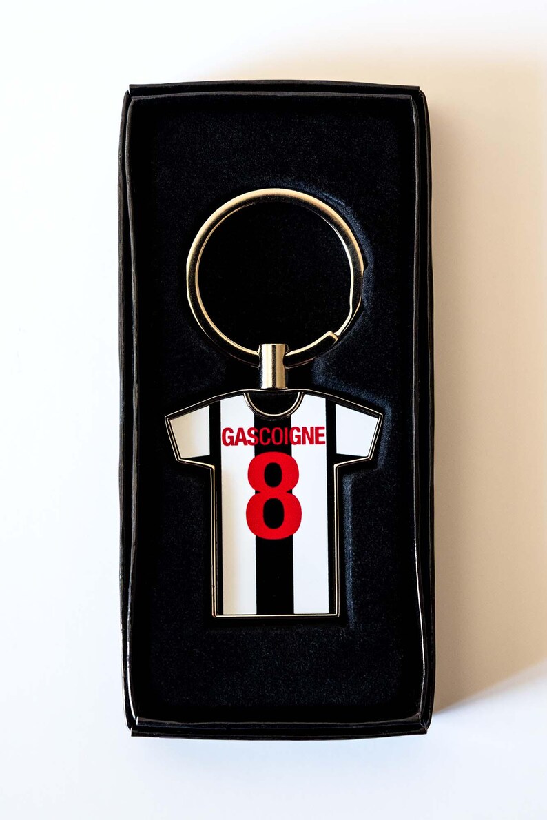 Puede incluir: Llavero con forma de camiseta de f&uacute;tbol a rayas blancas y negras con el nombre "GASCOIGNE" y el n&uacute;mero "8" en rojo. Presentado en una caja de regalo negra.