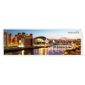 Puede incluir: Una vista panorámica nocturna de Newcastle, Inglaterra. El puente Tyne y el Sage Gateshead están iluminados, con el río Tyne en primer plano.