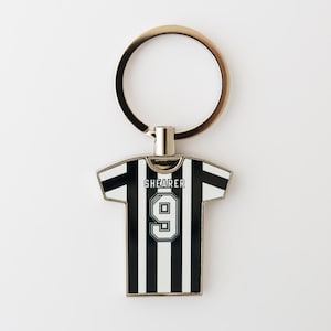 May include: A black and white striped soccer jersey keychain with the name "SHEARER" and the number "9" on the back. The keychain has a gold-colored ring attached to the top.