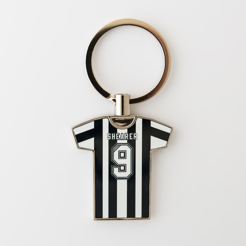 May include: A black and white striped soccer jersey keychain with the name "SHEARER" and the number "9" on the back. The keychain has a gold-colored ring attached to the top.