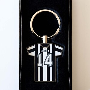 May include: Keyring featuring a black and white striped jersey design with the name "GINOLA" and the number "14". The keyring is attached to a silver-colored ring and presented in a black box.