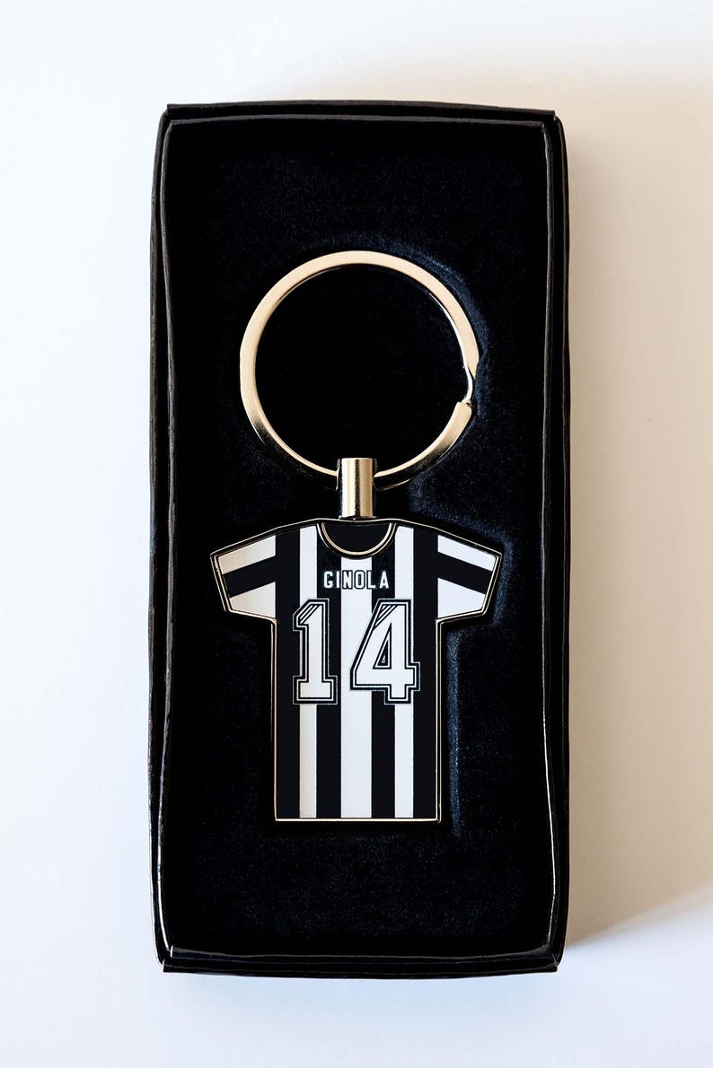 May include: Keyring featuring a black and white striped jersey design with the name "GINOLA" and the number "14". The keyring is attached to a silver-colored ring and presented in a black box.