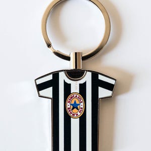 May include: A metal keychain featuring a black and white striped jersey design. The jersey has a round emblem with a blue star and the words "Newcastle Brown Ale". The keychain has a silver ring.