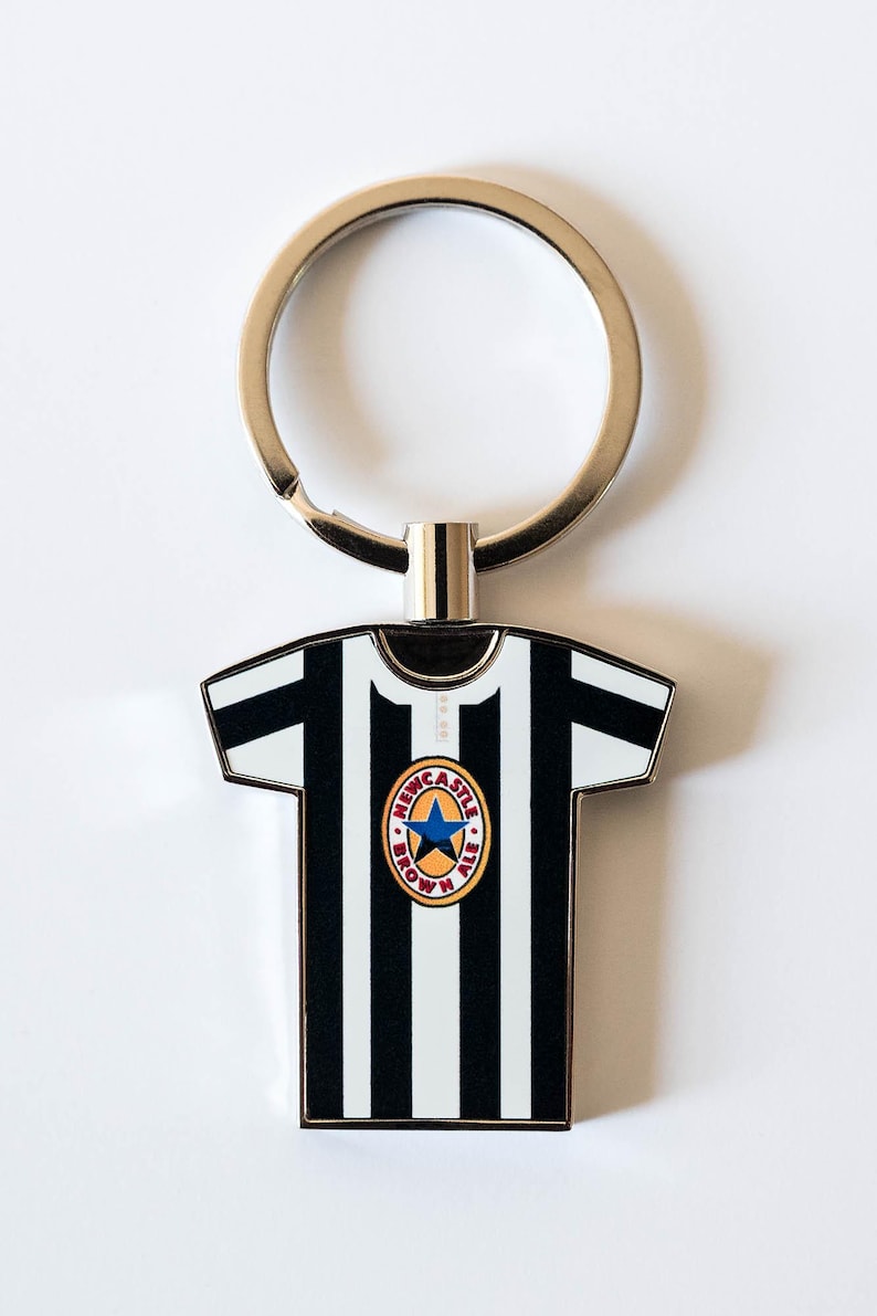 May include: A metal keychain featuring a black and white striped jersey design. The jersey has a round emblem with a blue star and the words "Newcastle Brown Ale". The keychain has a silver ring.