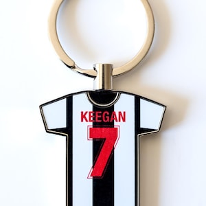 May include: Keyring shaped like a soccer jersey with black and white stripes, the name "KEEGAN" and the number "7" in red. The keyring has a silver-colored ring.