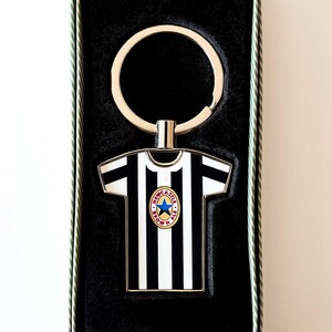 May include: A black and white striped keychain in the shape of a soccer jersey, with a team logo. The keychain is in a black box with a green and white striped border. The keychain has a silver key ring.