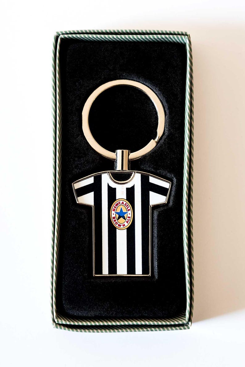 May include: A black and white striped keychain in the shape of a soccer jersey, with a team logo. The keychain is in a black box with a green and white striped border. The keychain has a silver key ring.