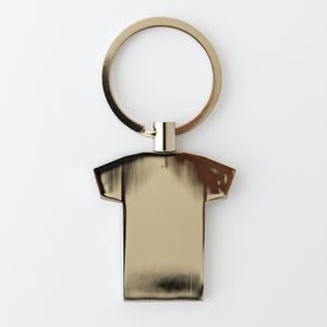 May include: A gold-colored metal keychain featuring a t-shirt shaped pendant. The pendant has a brushed metal finish and is attached to a circular key ring. The background is plain white.