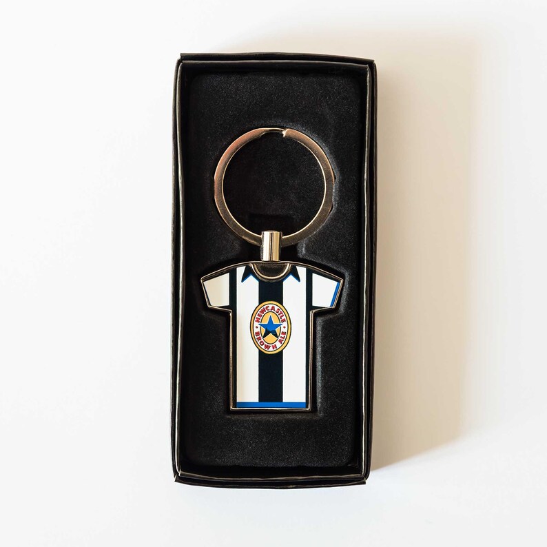 May include: A Newcastle United jersey-shaped keychain in a black gift box. The keychain features the team's black and white striped design, blue accents, and team logo. The keychain is made of metal and has a silver key ring.