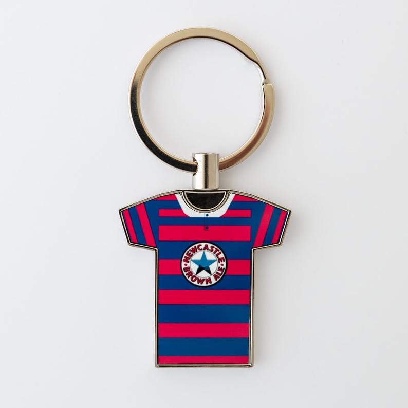 May include: A metal keychain featuring a miniature jersey design with red and blue stripes. The jersey has a white collar and a circular emblem that reads "Newcastle Brown Ale". The keychain has a gold-colored ring.