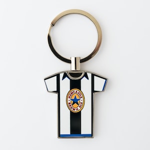 May include: A metal keychain featuring a miniature soccer jersey. The jersey is white with black vertical stripes and blue accents. The team logo is in the center. The keychain has a silver ring.