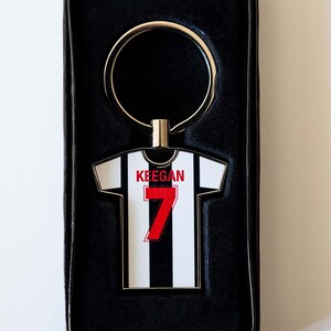 May include: A keychain in a black gift box. The keychain is shaped like a soccer jersey with black and white stripes, the number 7 in red, and the name "Keegan" above it. The keychain has a gold-colored ring.